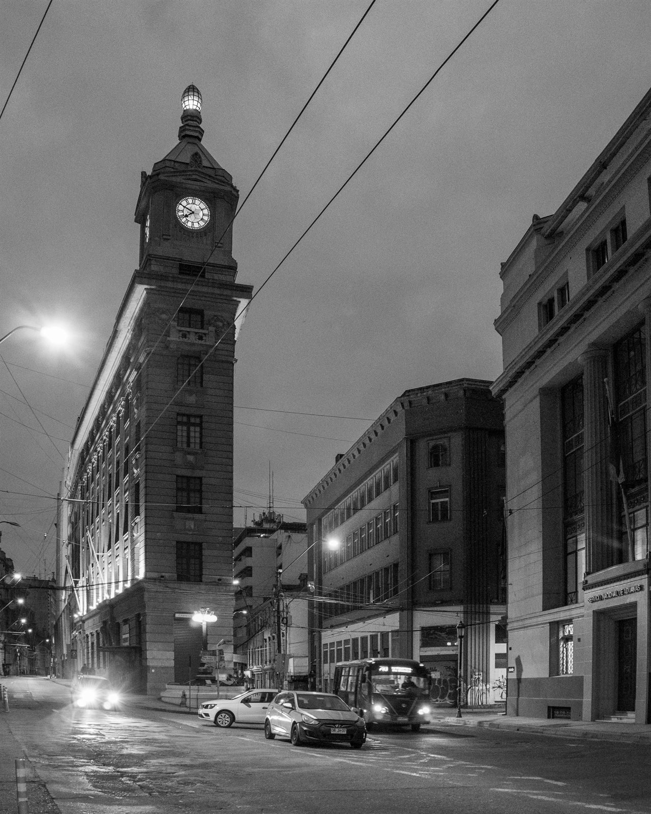 Edificio histórico con torre de reloj iluminada en una calle nocturna con tráfico y cables.