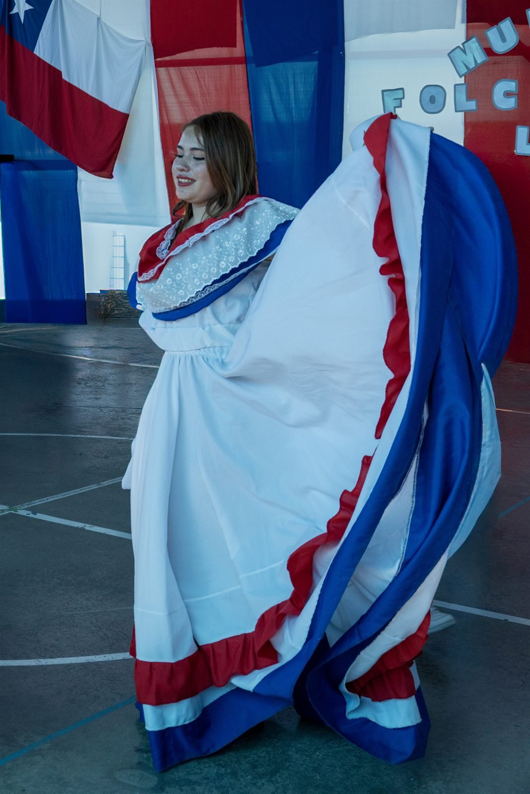 Mujer bailando la cueca con pañuelo blanco y rojo frente a una bandera chilena.