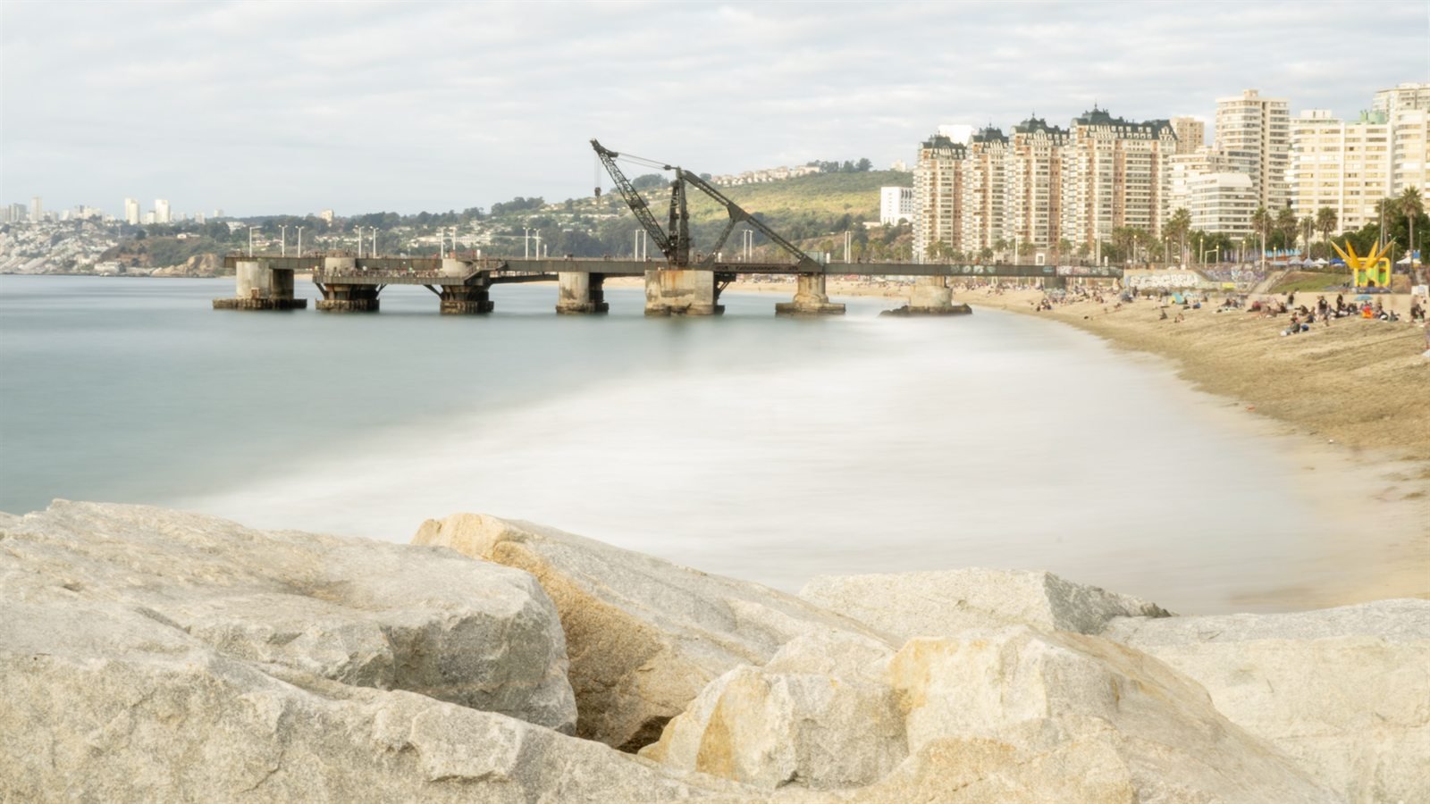 Muelle con grúa sobre el agua frente a una playa y edificios altos de ciudad.